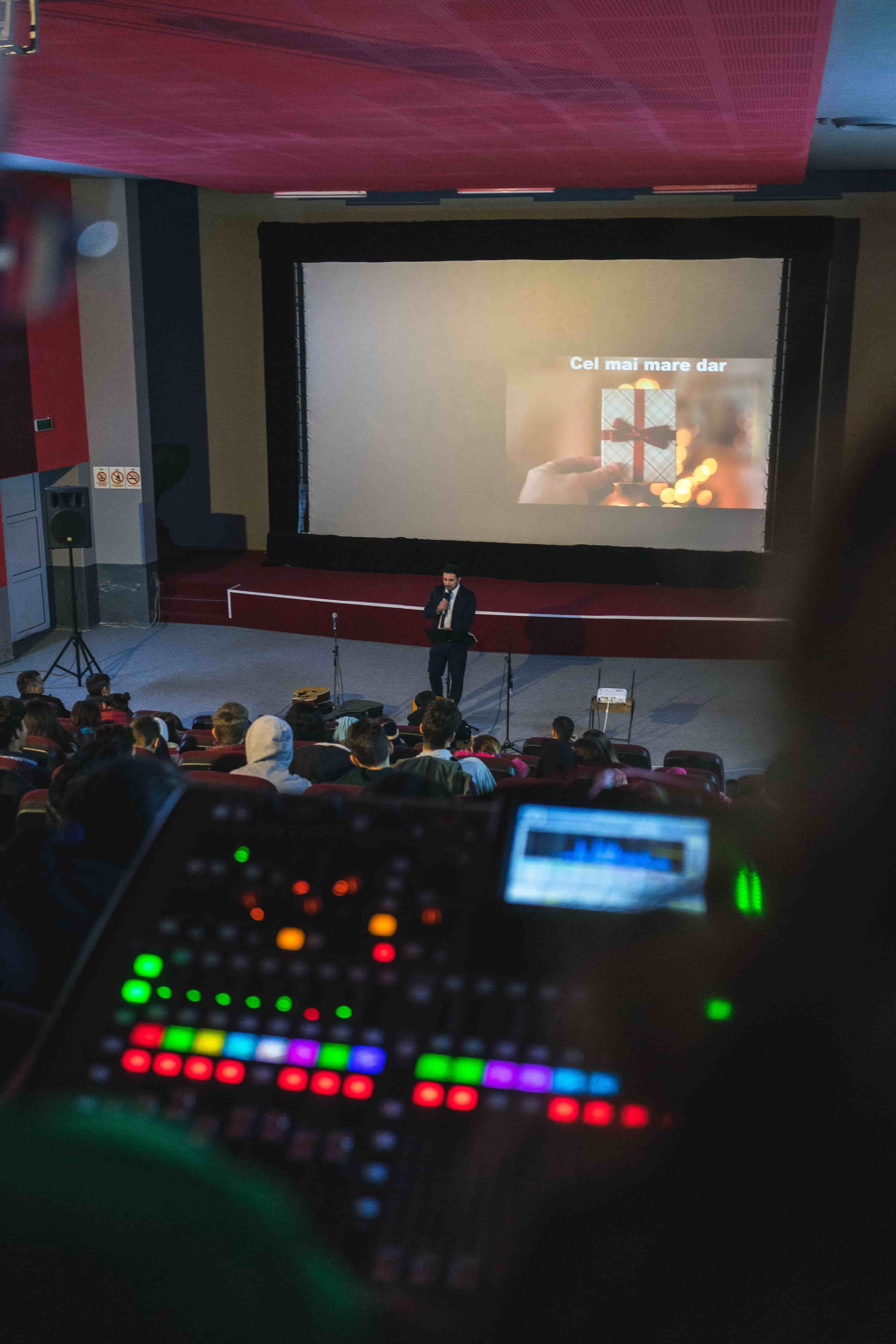 In der Mitte des Bildes ist ein Mann, der vor einer großen Leinwand etwas präsentiert. Im Vordergrund sieht man einen Mischer