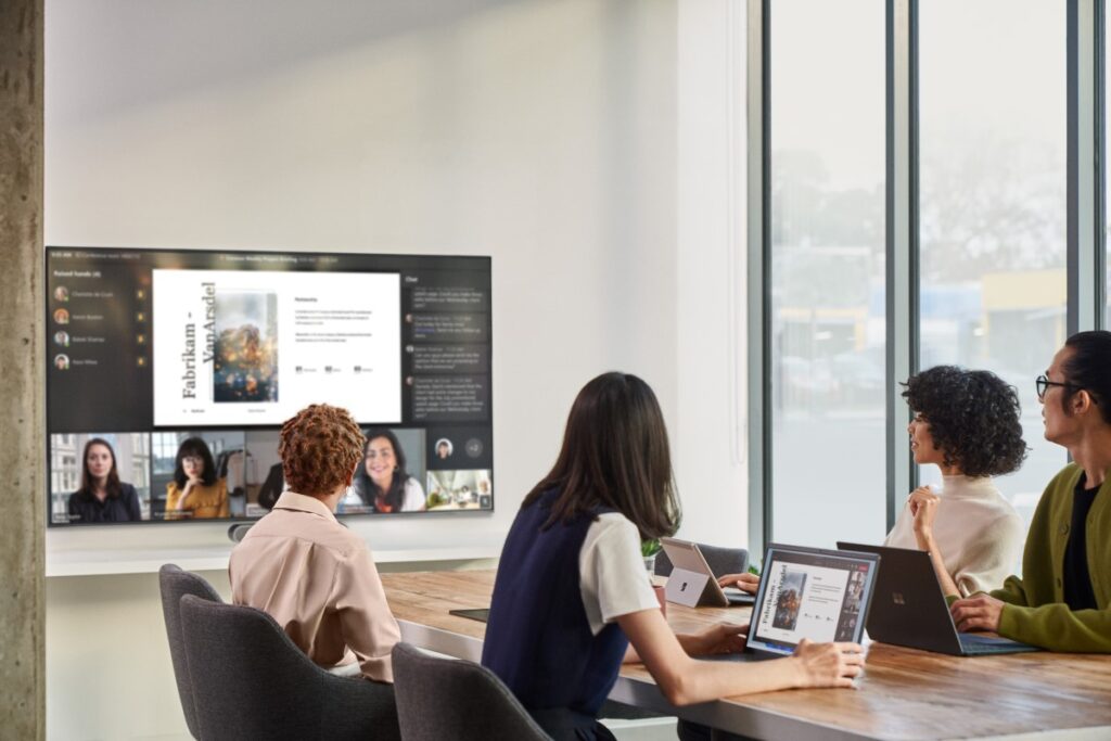 4 Personen in einem Konferenzraum sitzen an einem Tisch und kommunizieren mit 4 weiteren Personen die sich digital zugeschaltet haben.