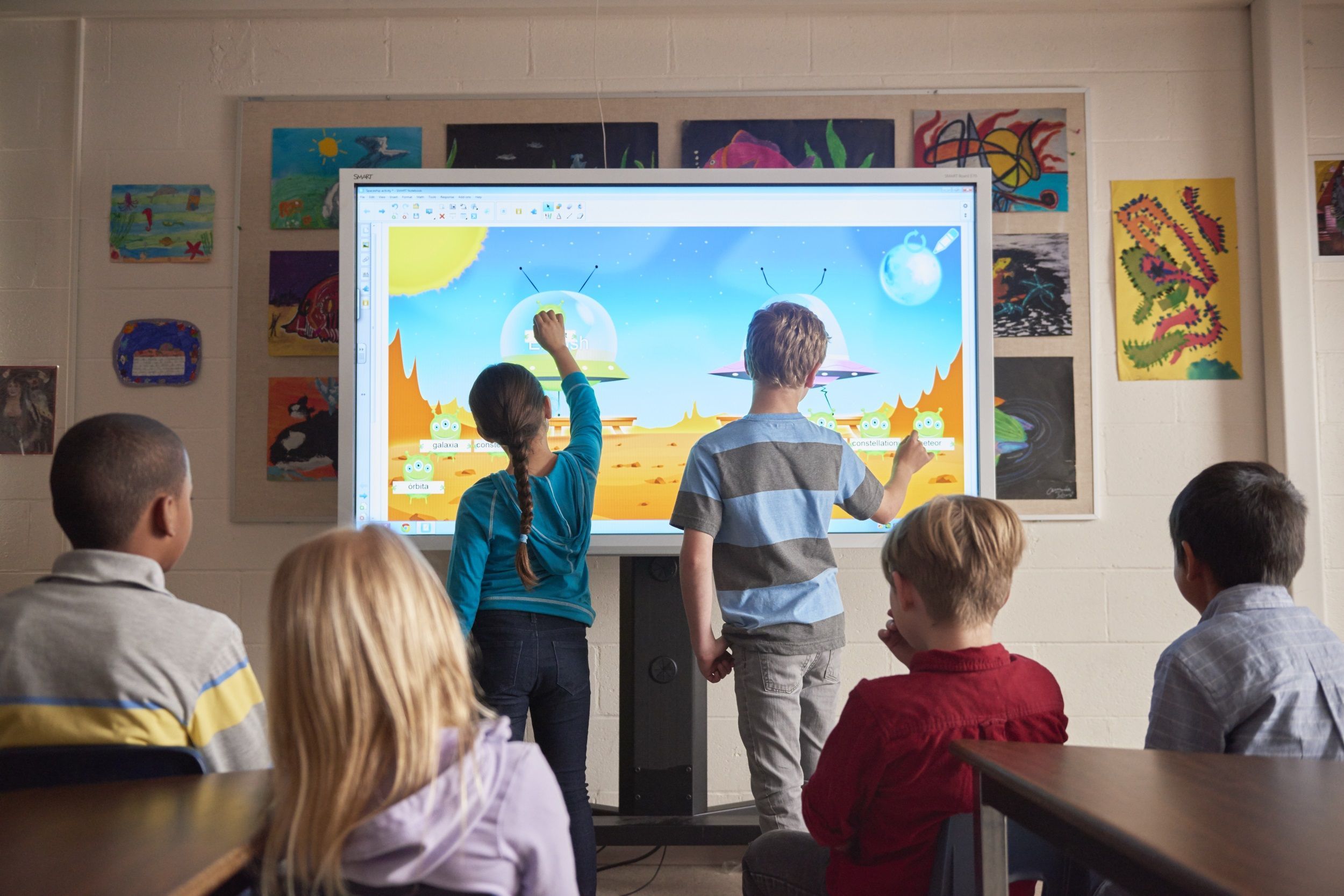 2 Kinder stehen vor einer digitalen Tafel und malen, während hinter ihnen 4 weitere Kinder sitzen und zuschauen.