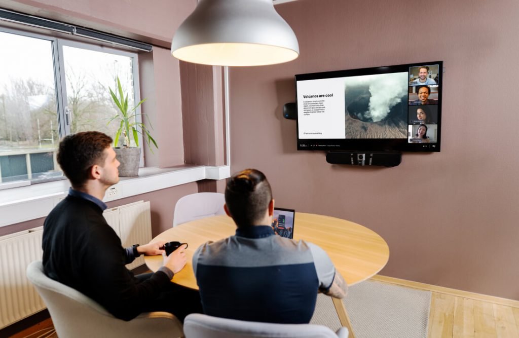 Zwei Personen sitzen an einem ovalen Tisch in einem Büro und betrachten einen Videokonferenzbildschirm, auf dem ein Vortrag mit mehreren Teilnehmern im Bild-in-Bild-Format läuft.