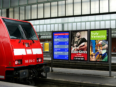 Auf dem Bild ist ein Bahnhof zu sehen. Links im Vordergrund steht ein roter Zug. Rechts im Hintergrund sind in einem Outdoor-Gehaeuse drei Displays im Hochformat installiert. Darauf werden Werbeinhalte und Informationen zu Abfahrtszeiten dargestellt.