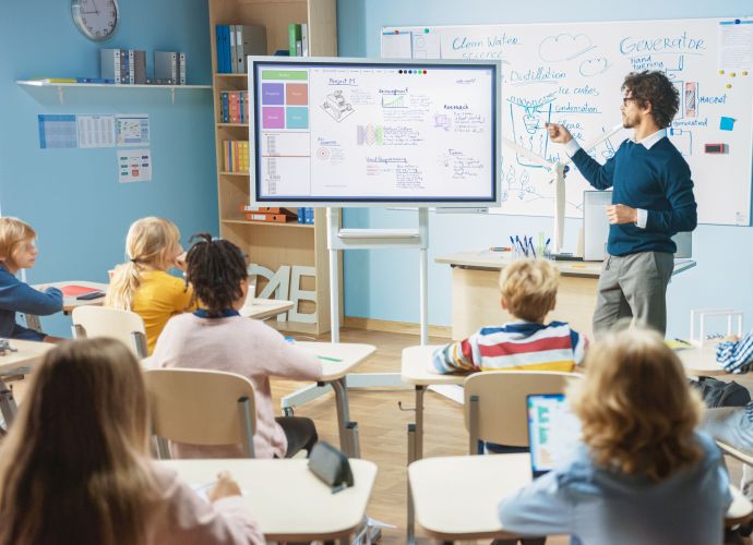 Das Bild zeigt eine Schulklasse, mit mehreren Kindern die aufmerksam auf eine Digitale Tafel schauen, auf die der Lehrer draufzeigt.