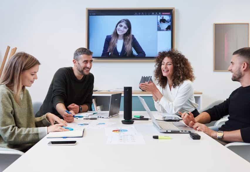 Vier Menschen sitzen an einem Tisch eines hellen Meetingraumes und halten ein hybrides Meeting ab. In der Mitte des Tisches steht eine Panasonic Konferenzkamera. An der Wand hängt ein Display.