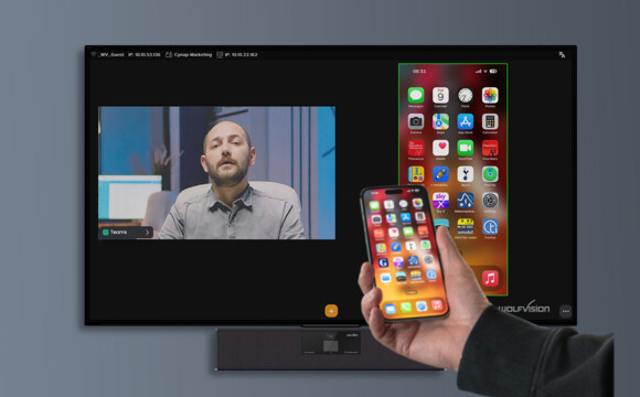 Eine Person hält ein Smartphone mit bunten App-Icons im Vordergrund, während im Hintergrund ein Mann auf einem großen Bildschirm zu sehen ist, der vermutlich Teil einer Videoübertragung ist.