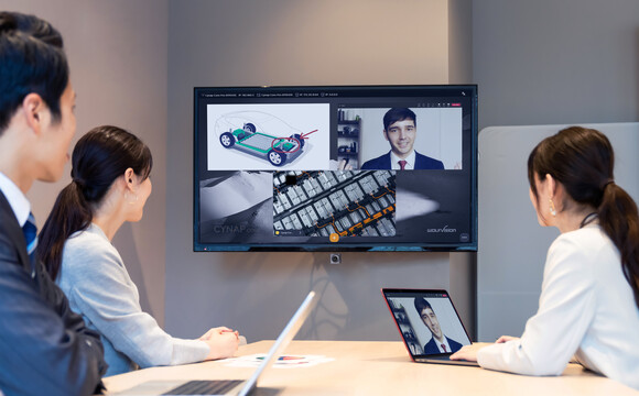 Drei Geschäftsleute führen in einem Büro eine Videokonferenz durch, bei der technische Zeichnungen und ein Teilnehmer auf einem großen Bildschirm dargestellt werden.