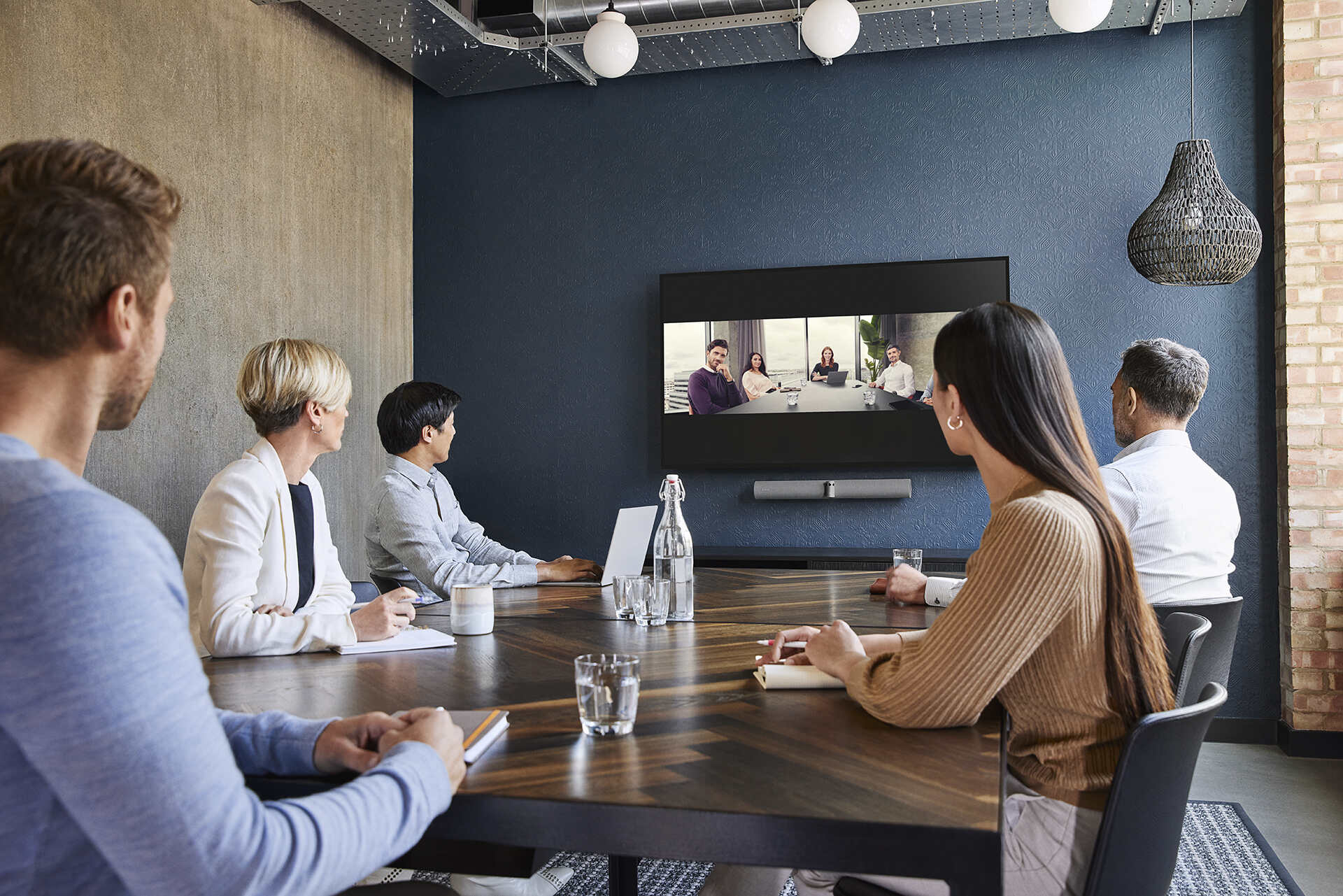 5 Personen sitzen in einem Videokonferenzraum und halten ein Meeting mit weiteren digitalen Teilnehmern, die auf einem Display angezeigt werden.