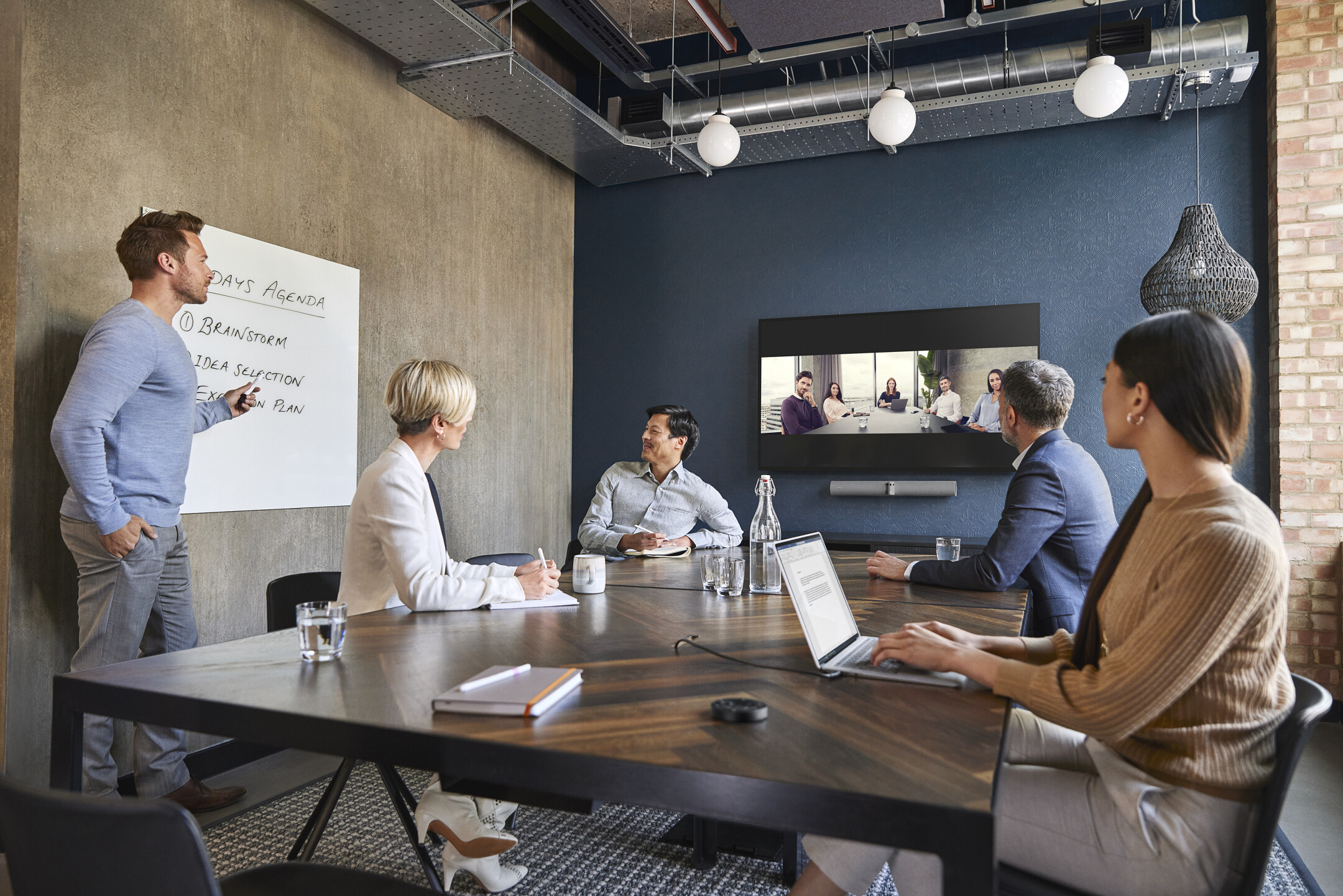 Auf dem Bild sind 5 Personen zu sehen, die sich in einem Videokonferenzraum befinden und sich in einem digitalen Meeting mit weiteren Kollegen unterhalten.