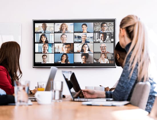 Mitarbeiter in einem Besprechungsraum schauen auf ein Display mit laufender Videokonferenz 