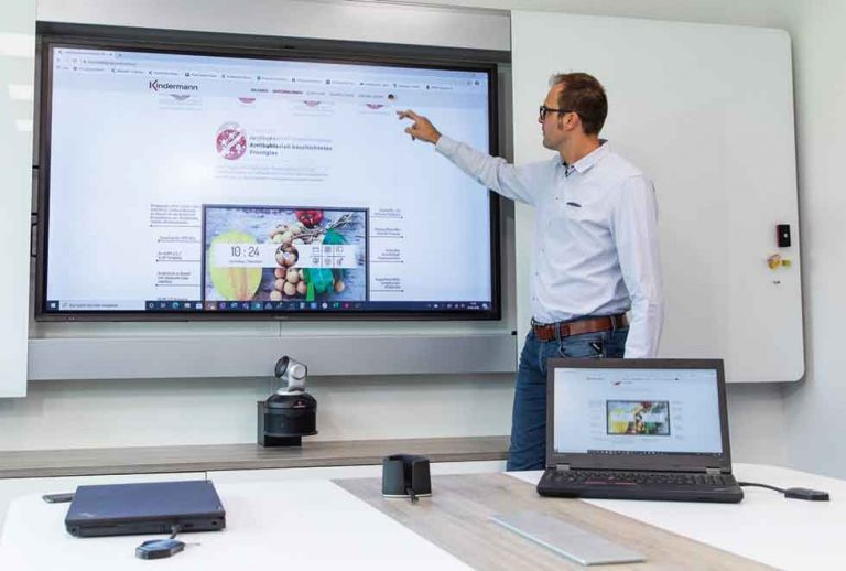 Ein Mann im Business-Casual-Look steht in einem Konferenzraum und zeigt auf einen interaktiven Whiteboard-Bildschirm, der eine Webseite anzeigt, neben einem aufgeklappten Laptop auf dem Konferenztisch.