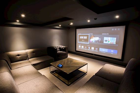 In einem dunklen Raum mit einem großen Sofa, hängt ein Optoma Projektor an der Decke und projiziert ein Bild auf die gegenüberstehende Wand.
