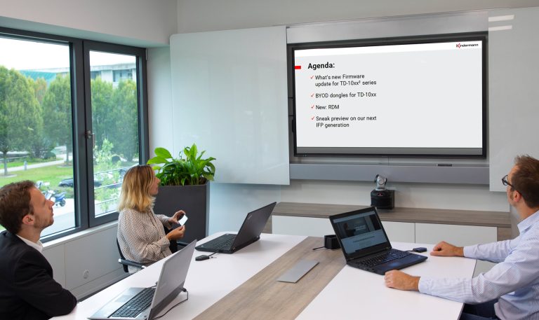 Drei Geschäftsleute sitzen in einem hellen Büro bei einem Meeting und blicken auf eine Präsentation, die auf einem großen Bildschirm angezeigt wird.