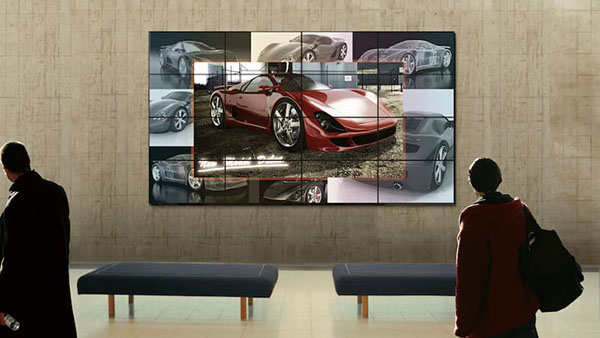 An der Wand im Hintergrund ist ein Panasonic Display installiert, auf dem Informationen und Bilder zu einem Auto angezeigt werden. Davor stehen zwei Baenke. Im Vordergrund stehen zwei Personen, die das Display betrachten