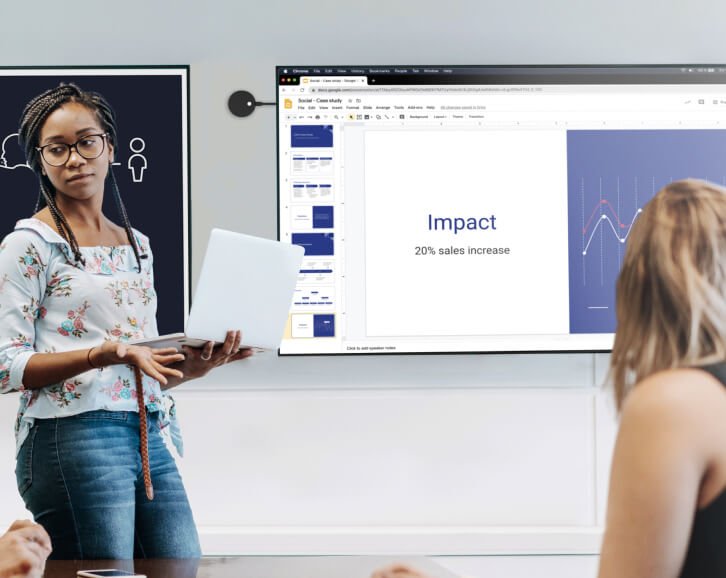 Eine Geschäftsfrau steht präsentierend vor einem Bildschirm mit einer offenen Powerpoint-Präsentation, die einen Punkt mit dem Titel "Impact" und eine Grafik einer 20%igen Umsatzsteigerung zeigt, während sie auf die Rückseite einer Kollegin blickt.