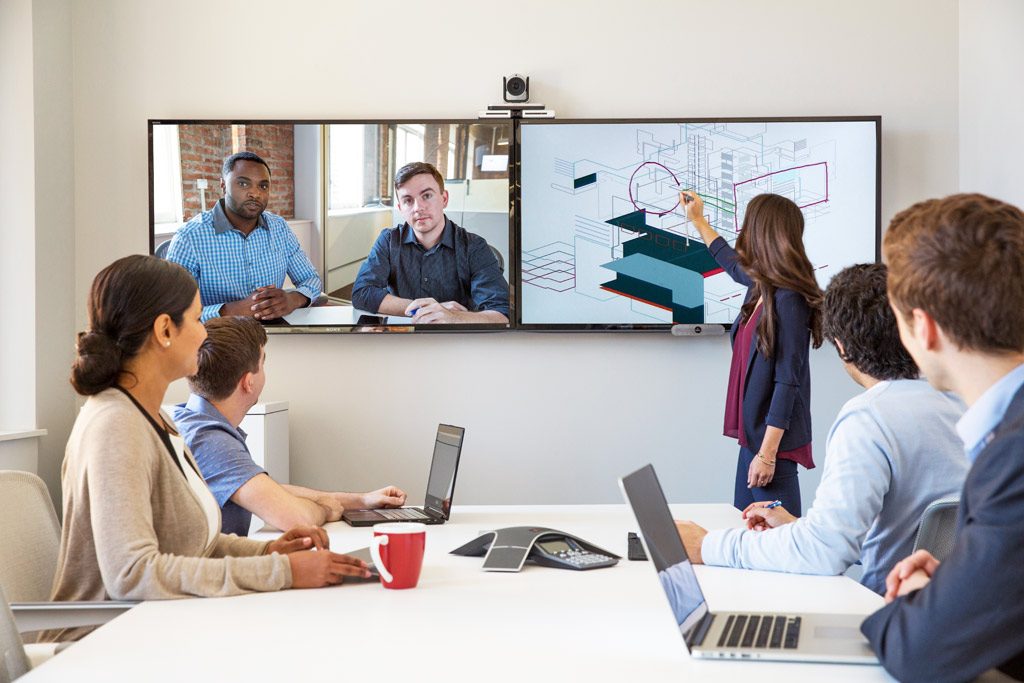 Eine Gruppe von Geschäftsleuten sitzt an einem Konferenztisch und interagiert mit Kollegen, die über eine große Bildschirmpräsentation zugeschaltet sind.